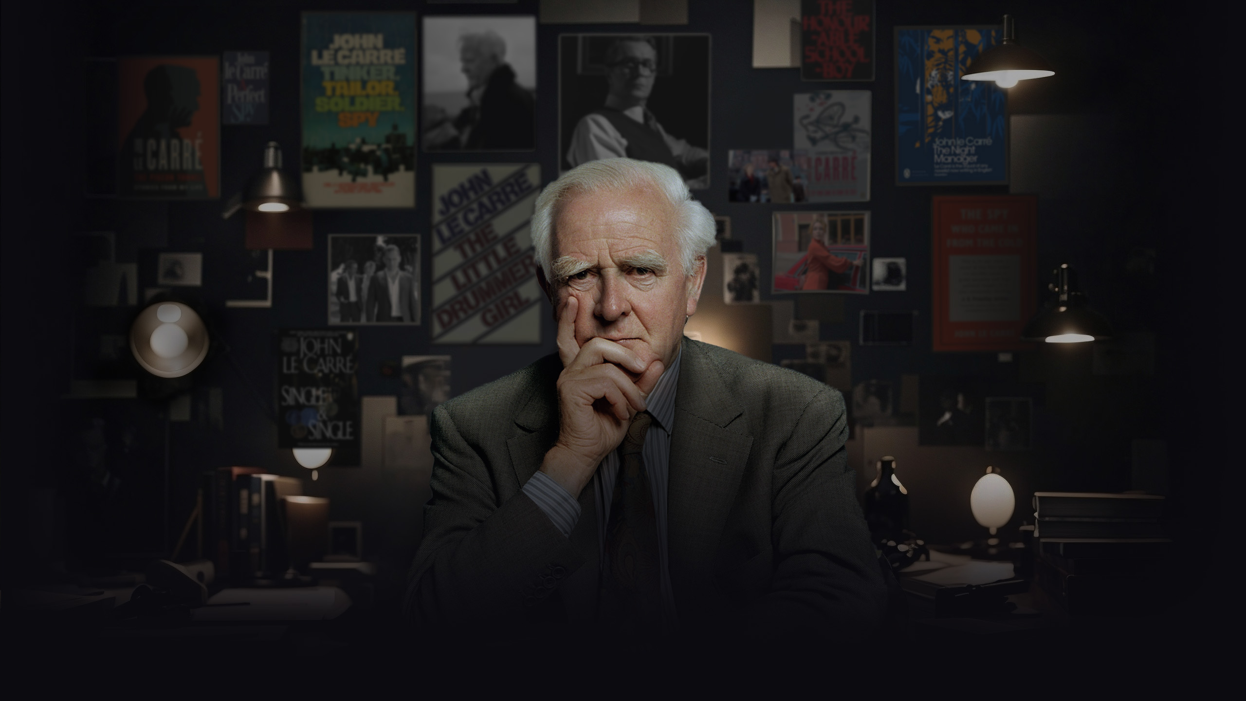 John Le Carré sitting in front of his desk with framed pictures of his books and films on the wall behind him.