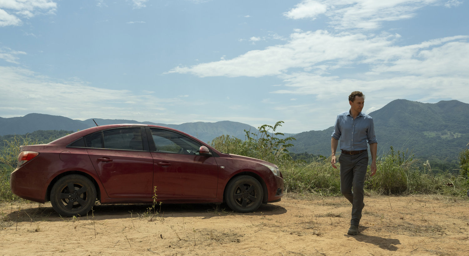 Man walking in front of red car with mountains in background