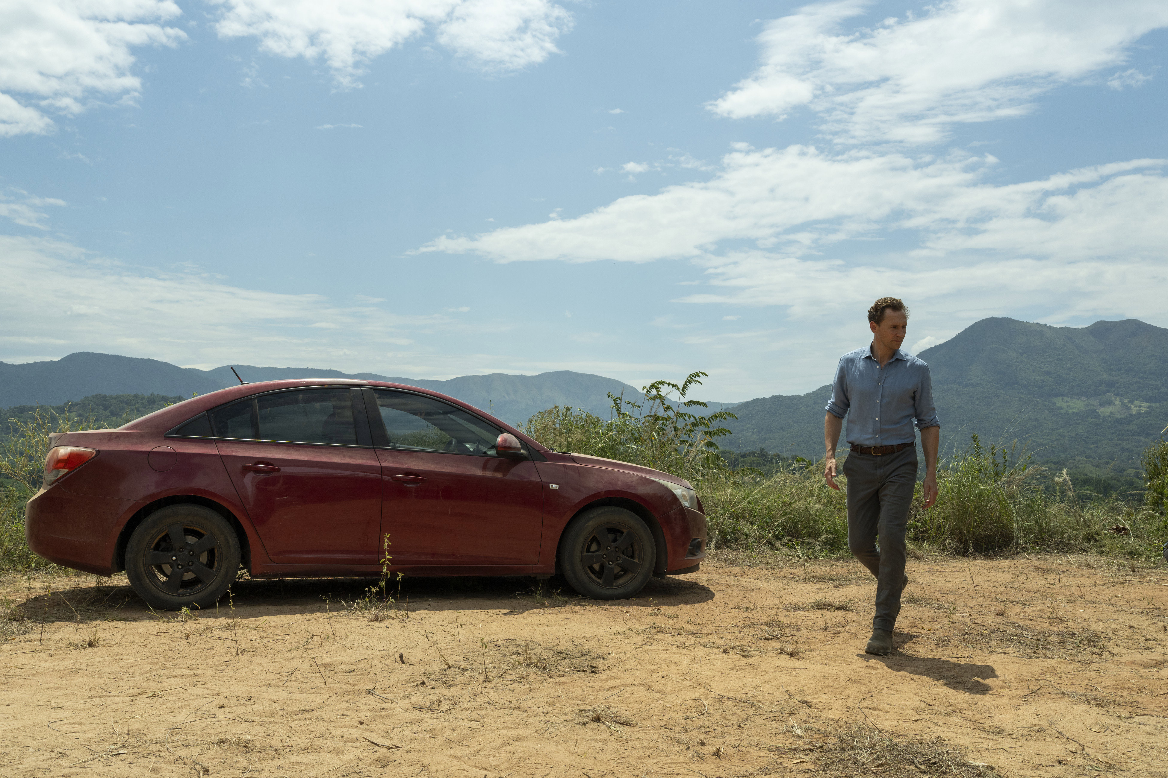 Man walking in front of red car with mountains in background